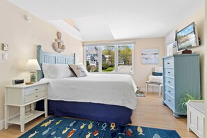 A bedroom with a bed, nightstand, dresser, and a large window with a view of houses.