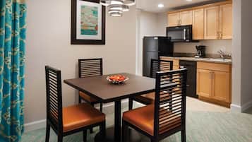 A dining area with a table, chairs, a bowl of fruit, a refrigerator, a microwave, and wooden cabinets.