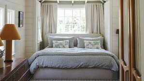 Una camera da letto con soffitto spiovente, un letto con lenzuola a quadri, una ventola a soffitto e una finestra con tende.