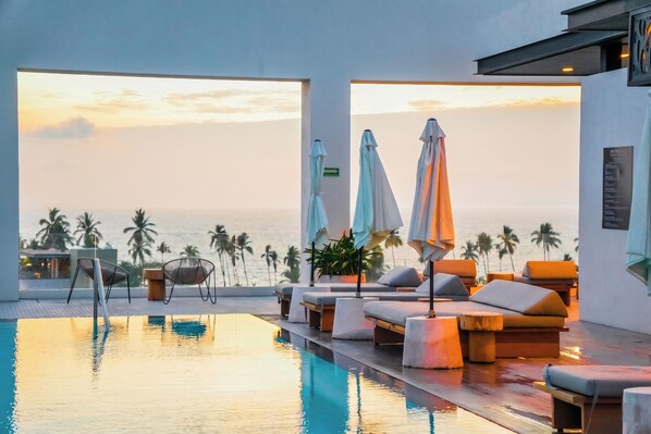 Ein unendlicher Pool mit Liegestühlen und Sonnenschirmen mit Blick auf einen Strand mit Palmen.