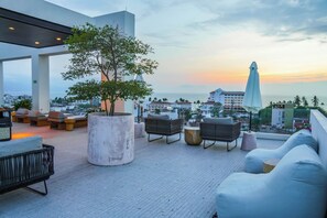 Eine Dachterrasse mit Sitzgelegenheiten, einem großen Pflanzgefäß und Blick auf Stadt und Meer bei Sonnenuntergang.