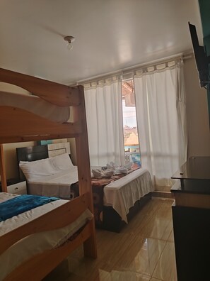 Ein Zimmer mit zwei Holzbetten, einem Schreibtisch, einem Deckenventilator und Blick auf die Stadt durch transparente Vorhänge.