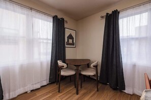 Una habitación con una mesa redonda de madera y dos sillas, un cuadro en la pared y cortinas negras en las ventanas.