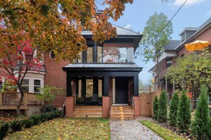 A two-story house with a front porch, a brick facade, and a small garden with trees and a wooden deck. - Luxury 4BR | High Park, Beach & Lake Views | Toronto (Toronto)