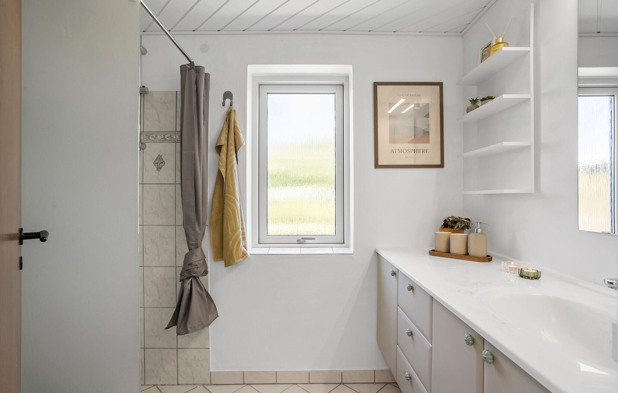 Une salle de bain avec une douche, une fenêtre, une œuvre d’art encadrée et un meuble-lavabo avec des articles de toilette.