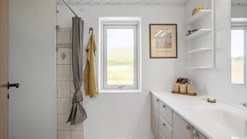 Une salle de bain avec une douche, une fenêtre, une œuvre d’art encadrée et un meuble-lavabo avec des articles de toilette.