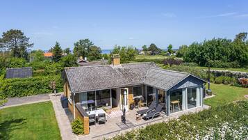 Une maison moderne dotée d’une grande extension en verre, d’un patio avec mobilier d’extérieur et d’une vue sur la mer.