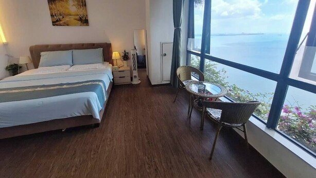 A bedroom with a bed, bedside table, lamp, and a view of the sea through large windows.