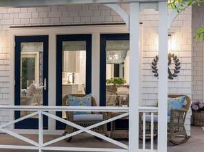 A covered porch with wicker furniture, a white railing, and a brick wall. - Bald Head Island retreat, where coastal elegance meets relaxed island living (Bald Head Island)
