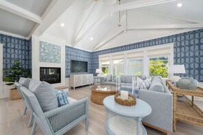 A spacious living room with a fireplace, large windows, and a ceiling with exposed beams. - Bald Head Island retreat, where coastal elegance meets relaxed island living (Bald Head Island)
