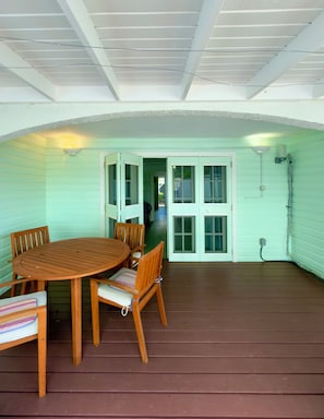 Een overdekt terras met een houten tafel en stoelen, een uitzicht op het huis door open deuren, en een plafond met zichtbare balken.