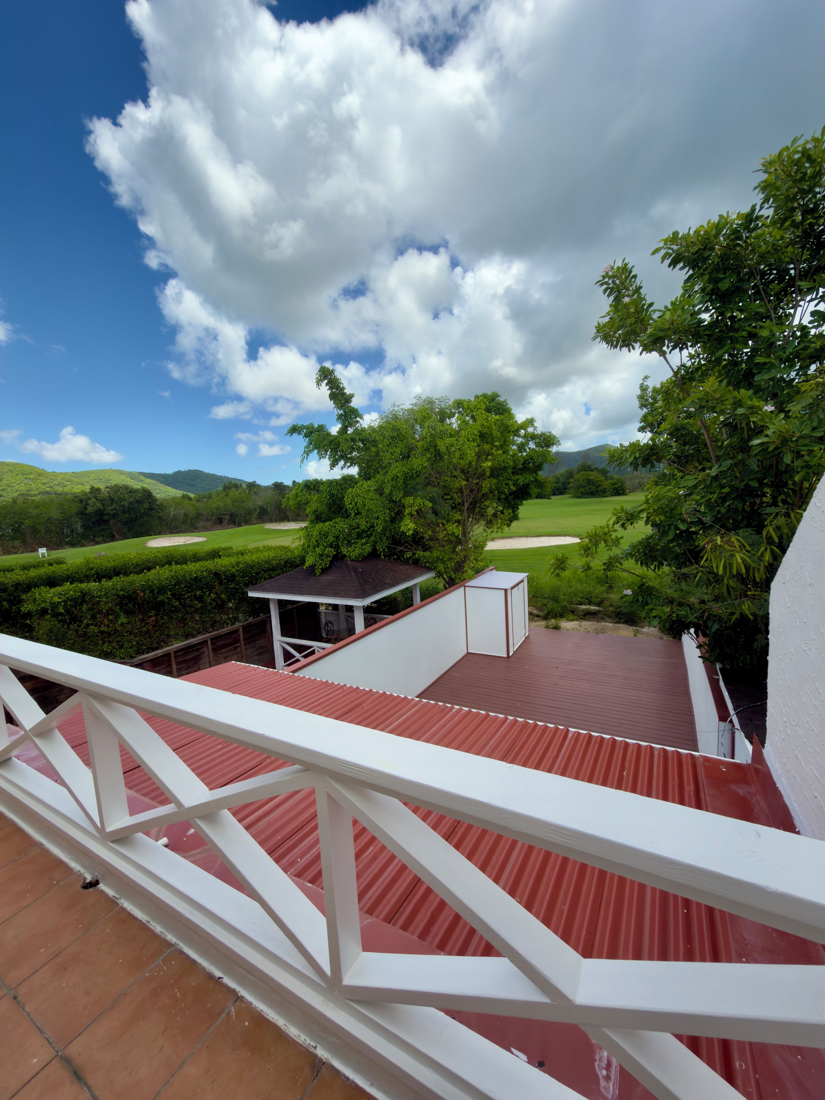 A terrace with a red roof, white railings, and a view of a green landscape with hills and a clear sky.