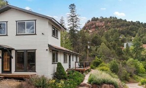 Ein zweigeschossiges Haus mit einer Terrasse, umgeben von Grünpflanzen und einer Hügelsicht.