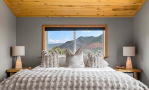 Ein Schlafzimmer mit einem großen Bett, einem Fenster mit Bergblick, zwei Nachttischlampen und einer Holzbalkendecke.