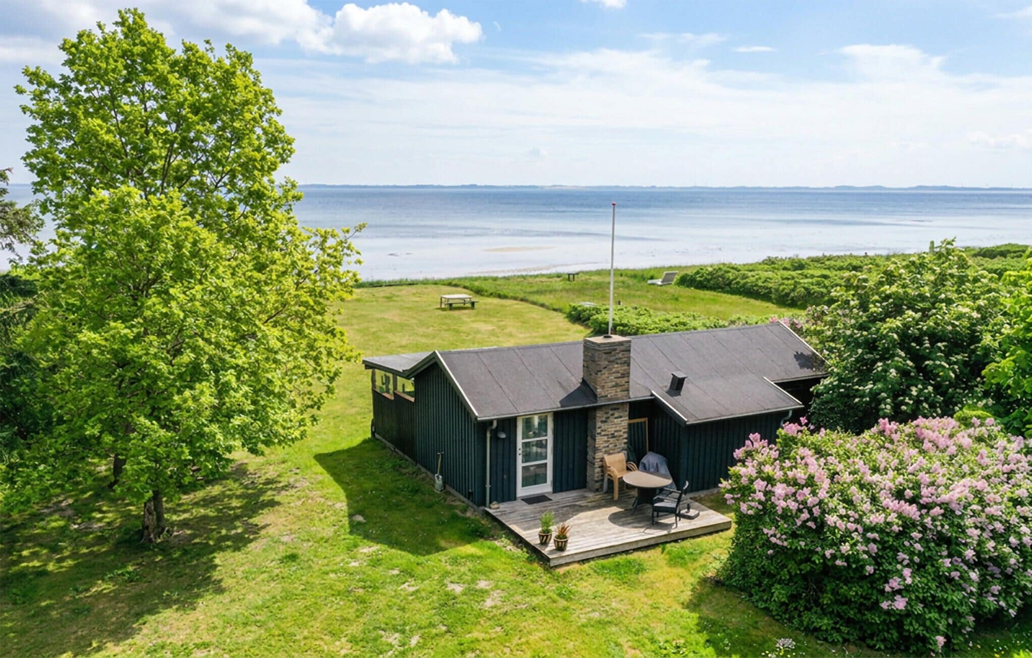 A modern house with a stone chimney, a deck with outdoor furniture, and a view of the sea.