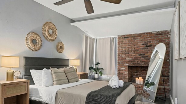 A modern bedroom with a brick fireplace, a large bed with a headboard, bedside lamps, and decorative wall art. - Serene 4-bedroom house with AC in charming Alhambra (Alhambra)