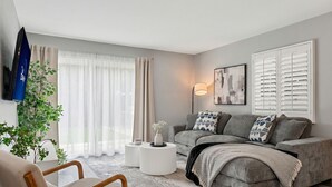 A modern living room with a grey sofa, a wooden chair, a round coffee table, and a large window with sheer curtains. - Serene 4-bedroom house with AC in charming Alhambra (Alhambra)