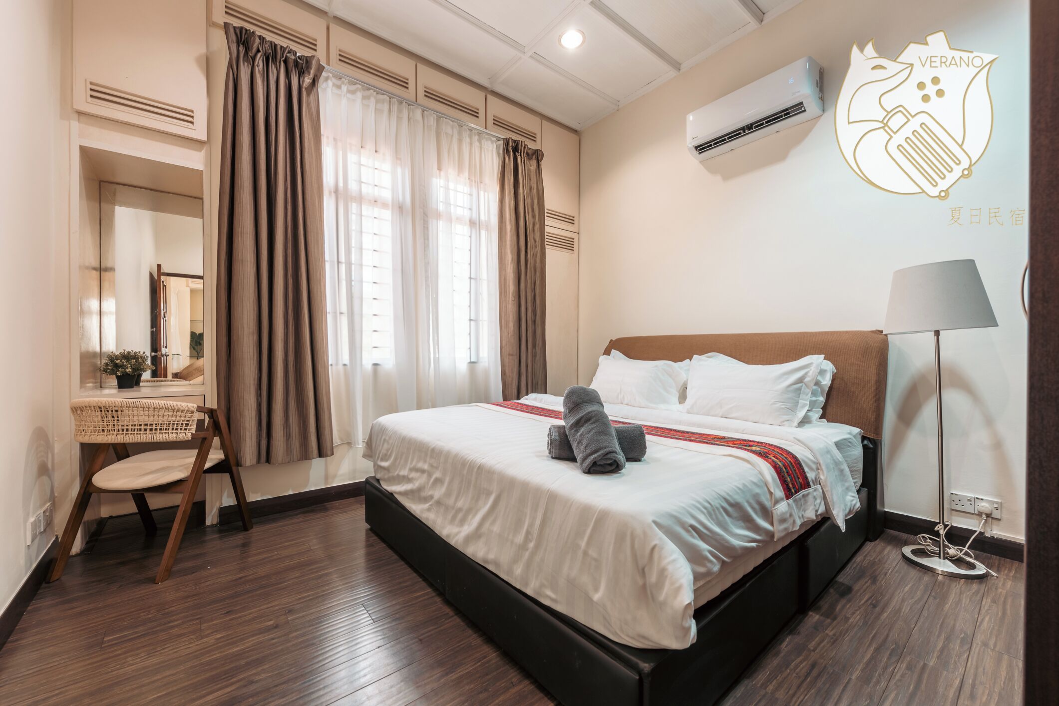 A hotel room with a bed, a desk, a chair, a lamp, and an air conditioning unit.