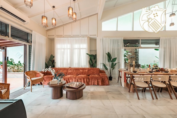 A spacious living area with a sofa, dining table, and chairs, large windows, and hanging lights.