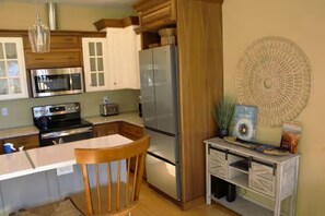 A kitchen with wooden cabinets, a stainless steel refrigerator, a microwave, and a dining area with a wooden chair. - The Blue House on Barr -Charming Cottage Retreat in the Heart of Canon City (Cañon City)