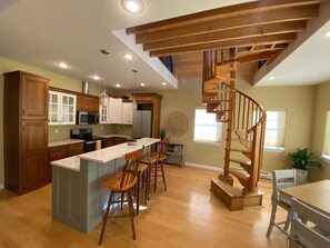 A kitchen with wooden cabinets, a central island, bar stools, a spiral staircase, and a dining area. - The Blue House on Barr -Charming Cottage Retreat in the Heart of Canon City (Cañon City)