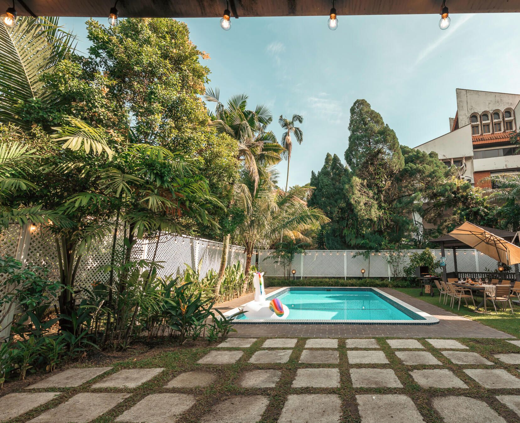 A swimming pool with a tiled deck, surrounded by lush greenery and a white fence.