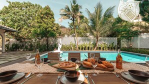 A dining table set with plates, cutlery, glasses, and food items, with a pool and lush greenery in the background.