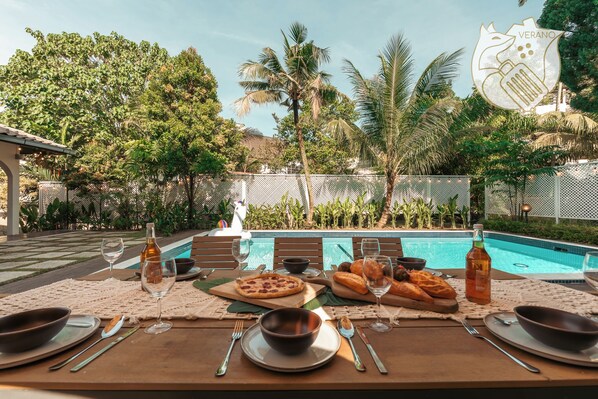 A dining table set with plates, cutlery, glasses, and food items, with a pool and lush greenery in the background.