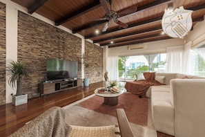 A living room with a stone wall, wooden ceiling beams, a ceiling fan, a TV, a sofa, a coffee table, and a view of the outdoors.