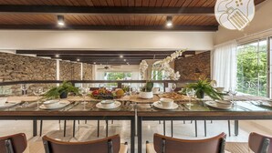 A dining area with a long table set for four, surrounded by chairs, with a stone wall and a view of greenery outside.
