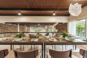 A dining area with a long table set for four, surrounded by chairs, with a stone wall and a view of greenery outside. - 42Pax 9BR Damansara Pool Villa by Verano (Kuala Lumpur)