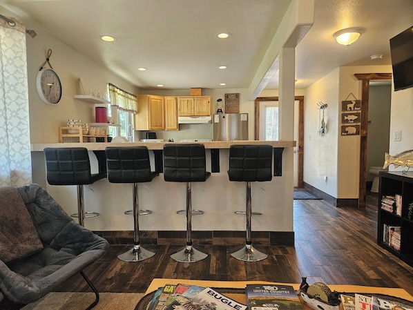 A modern kitchen with wooden cabinets, a central island with bar stools, a dining area with a grey chair, and a bookshelf with magazines.