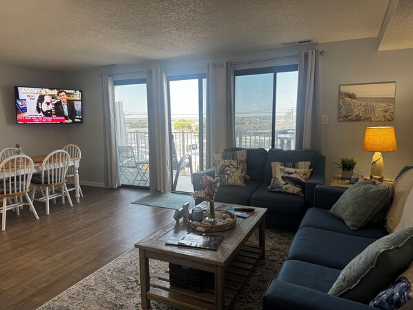Ein Wohnzimmer mit Fernseher, Essbereich mit Stühlen, einer Couch, einem Couchtisch und Blick durch große Fenster.