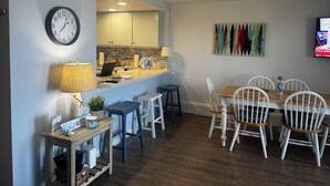 Un comedor con una mesa de madera y sillas blancas, una cocina al fondo, un reloj en la pared y un televisor montado en la pared.