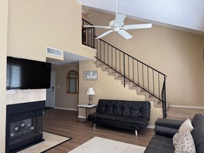 A living room with a fireplace, a black leather sofa, a black leather couch, a white ceiling fan, and a staircase. - Spacious St. George Condo with Loft, Sleeps 10 (St. George)