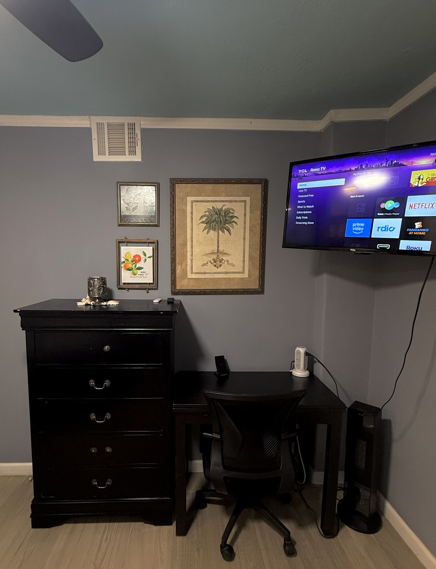 Une pièce avec un bureau noir, une chaise, un téléviseur fixé au mur et un tableau représentant un arbre.