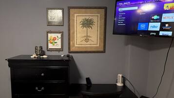 Une pièce avec un bureau noir, une chaise, un téléviseur fixé au mur et un tableau représentant un arbre.