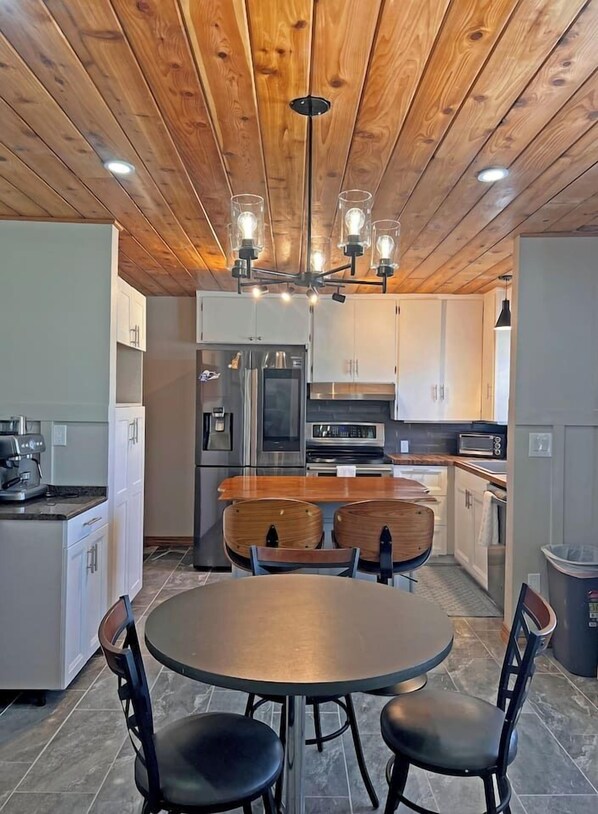 A modern kitchen with wooden ceiling, stainless steel appliances, a round dining table, and two chairs. - Omaha Estate·4BR·Hot Tub·Sauna·Game Room·Sleeps13 (Omaha)