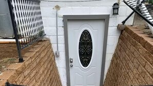 A set of concrete steps leading to a white door with a metal grille, flanked by brick walls and metal handrails.