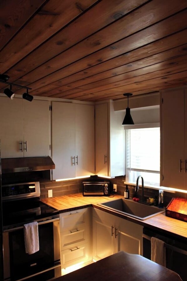A kitchen with wooden ceiling, white cabinets, a black stove, and a window with blinds. - Omaha Estate·4BR·Hot Tub·Sauna·Game Room·Sleeps13 (Omaha)