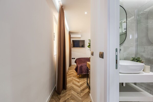 A hotel room with a herringbone floor, a bed with a purple bedspread, a mounted TV, and a bathroom with a glass shower enclosure. - Konak Lemon Guesthouse Heart of Vlora (Vlorë)