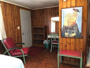 Une pièce aux murs en bois, avec une porte, une chaise rouge, une chaise verte, une petite table et une affiche accrochée au mur.