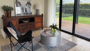 A modern living room with a black leather chair, a wooden cabinet with a framed picture, a round grey ottoman with a plant, and a view of a garden through large glass doors. - L&L Golfside Retreat (Benowa)