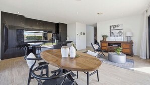 A modern kitchen with dark cabinetry, a dining area with a wooden table and chairs, and a view of the outdoors. - L&L Golfside Retreat (Benowa)