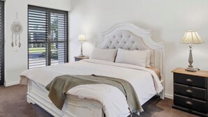 A bedroom with a white bed, two bedside tables, a nightstand, a window with blinds, and a dreamcatcher on the wall. - L&L Golfside Retreat (Benowa)