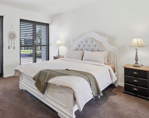A bedroom with a white bed, two bedside tables, a nightstand, a window with blinds, and a dreamcatcher on the wall.