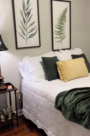 A bed with a white comforter, multiple pillows, a dark green throw blanket, and two framed botanical prints on the wall.