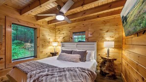 Une chambre dans une cabane en bois, avec un lit, un ventilateur de plafond et deux fenêtres.