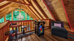 Une pièce mansardée avec un plafond en bois, un lustre, une petite table avec des chaises, un canapé et une borne d’arcade aux couleurs vives.