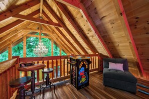 Ein Dachzimmer mit Holzbalkendecke, einer Chandelier, einem kleinen Tisch mit Stühlen, einem Sofa und einem farbenfrohen Spielautomaten.